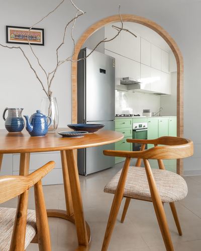 Round wooden dining table with wishbone chairs framed by an arched doorway leading to a sage green kitchen