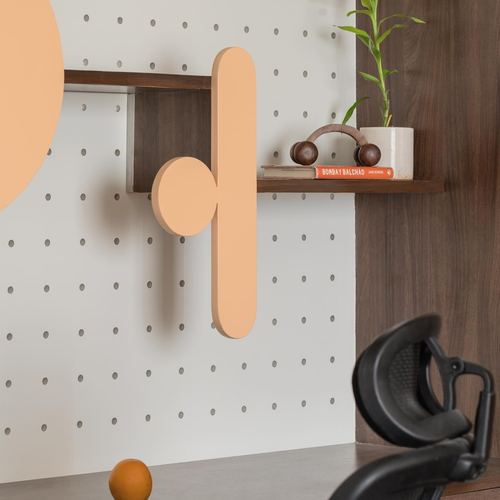 Pegboard wall with wooden shelf, sculptural orange forms, plant, and book beside a desk chair