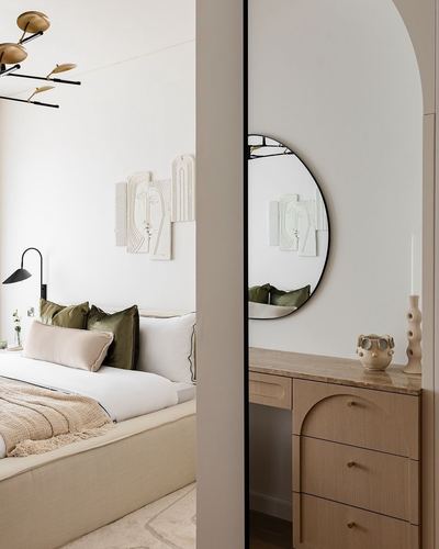 Neutral bedroom with olive cushions seen through an arched opening beside a round mirror and wood vanity