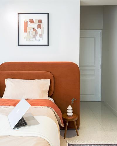 Bedroom with terracotta curved velvet headboard, cream linens, laptop, and wooden side table.