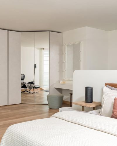 Minimalist bedroom corner with mirrored wardrobe, lit vanity mirror, and wood-toned nightstand beside white bed