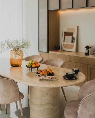 Oval travertine dining table with pink velvet chairs, fruit bowl and croissants, cabinetry behind