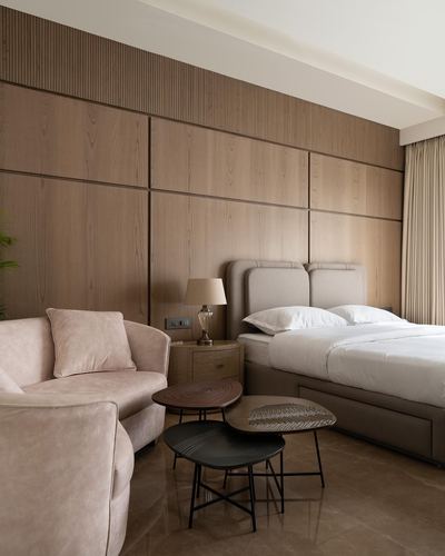 Bedroom with wood-panelled wall, beige upholstered bed, blush armchair and layered round coffee tables.