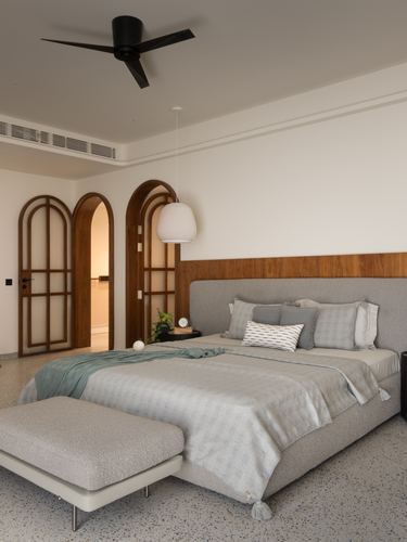 Bedroom with grey upholstered bed, walnut headboard panel, terrazzo floor, and two arched wooden doors