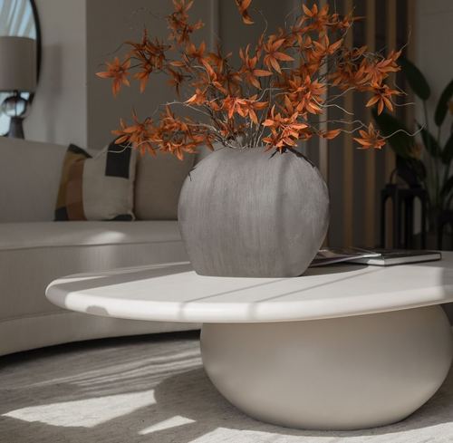 Grey textured vase with orange dried branches on a round stone-look coffee table beside a beige sofa