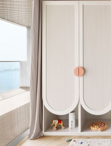 Ribbed white wardrobe with arched base and pink circular handle beside window overlooking sea, children's toys below