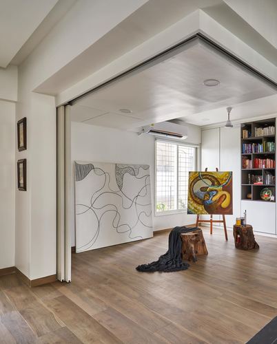 Open studio space with wooden floor, easel painting, tree-stump stools, and bookshelf under a folded ceiling beam