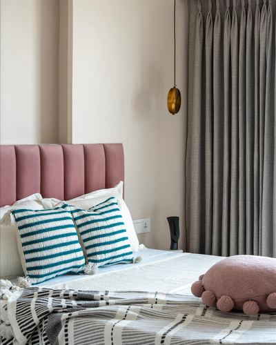 Pink channel-tufted headboard with striped cushions beside a brass pendant light and grey curtain