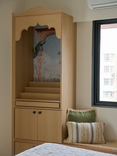 Built-in wooden pooja unit with carved arch and peacock mural beside a cushioned window seat