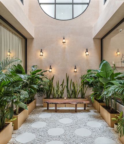 Double-height courtyard with plaster walls, lantern sconces, round skylight, live-edge bench, and dense planting on a pebble floor