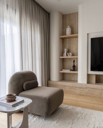 Curved beige armchair beside a stone niche with floating wood shelves and sheer curtains