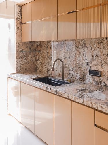 Kitchen counter with veined stone backsplash, black sink, brass faucet, and fluted tan cabinetry