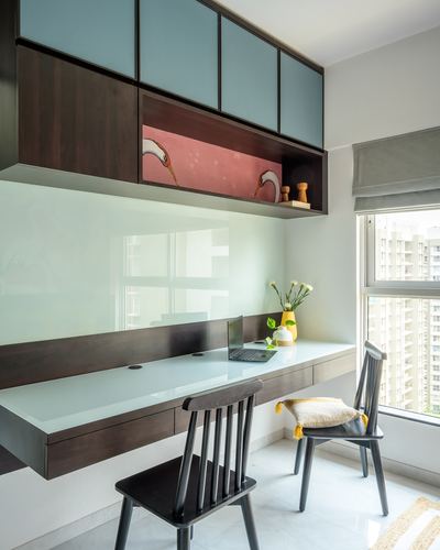 Floating wood desk with glass top below blue cabinets and pink patterned niche, window view