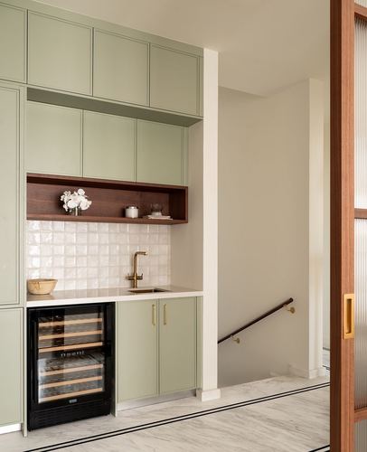 Sage green butler's pantry with tiled backsplash, brass faucet, wine fridge, and staircase beyond