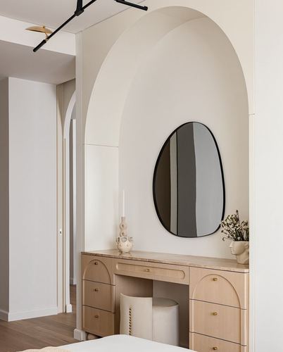 Arched niche with organic mirror above a wooden vanity desk and drum stool in a beige bedroom