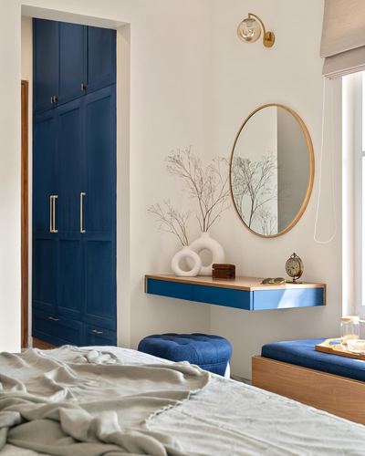 Bedroom corner with navy wardrobe, floating vanity, round mirror, and tufted ottoman