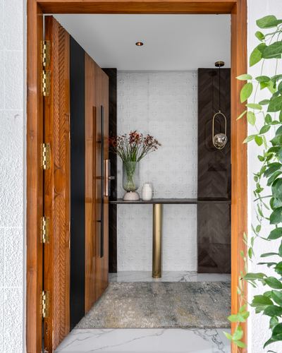 Open carved wooden front door revealing a foyer with textured tile wall, brass-legged console, and vase of branches