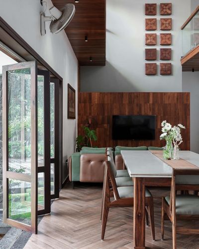 Dining table beside sage sofa under double-height wood wall with framed tile art, rainy garden view through glass doors