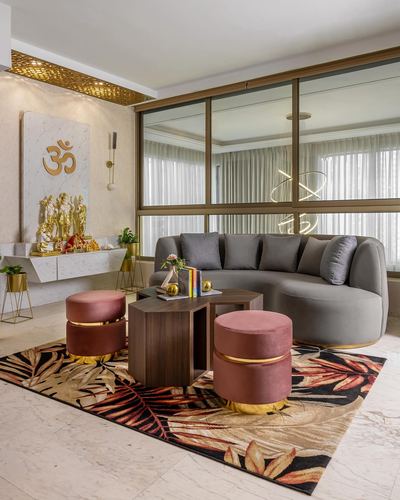 Living room with curved grey sofa, pink ottomans, floral rug, and pooja niche with om symbol