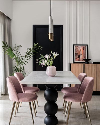 Dining table with marble top on black sculptural base, surrounded by pink upholstered chairs beside a black door