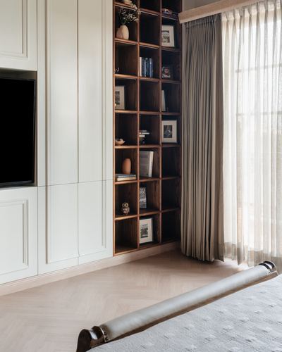 Bedroom corner with wooden grid bookshelf, cream panelled cabinetry, and sheer curtained window