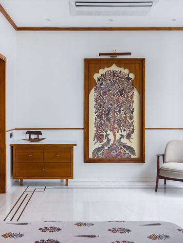 Bedroom wall with arched wooden frame holding pichwai tree-of-life painting above chest of drawers and armchair