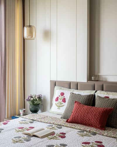 Bed with floral quilted linens and cushions against a paneled wall, amber pendant light beside