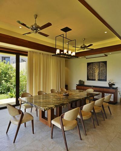 Long stone-top dining table with twelve chairs under a linear chandelier, glass wall opens to garden