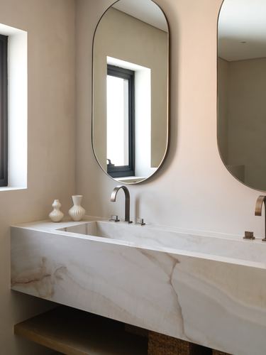 Floating veined stone sink beneath two oval mirrors and a window in a beige plaster bathroom