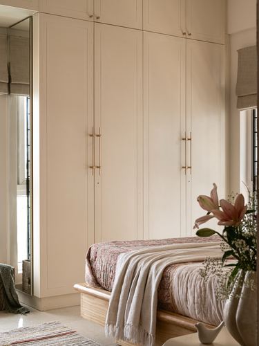Cream wardrobe wall with brass handles beside a bed with patterned throw and lilies