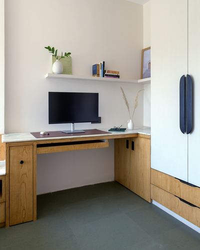 Wooden corner desk with monitor beneath a floating shelf, beside a tall wardrobe