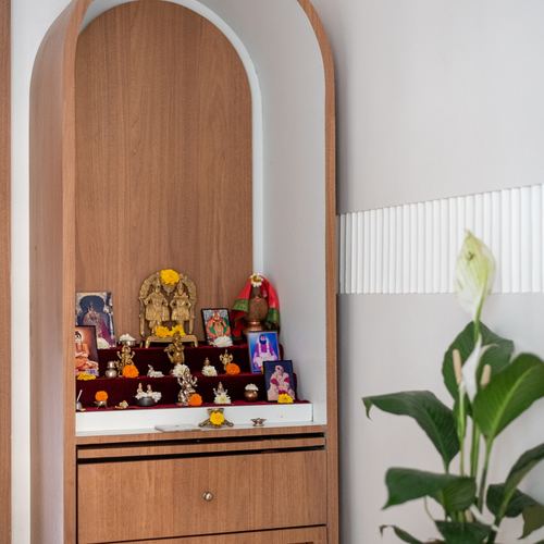 Arched wooden pooja cabinet with deity idols beside a fluted white wall and plant