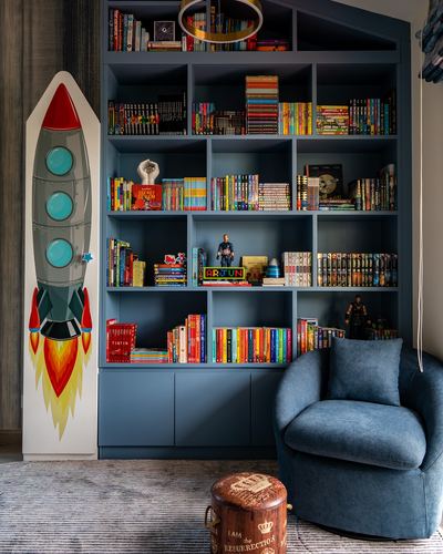 Blue built-in bookshelf filled with colorful books beside a rocket-shaped wall decor and blue armchair