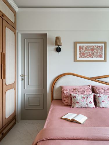 Bedroom corner with pink bedding, curved wood headboard, cane wardrobe, and framed floral art