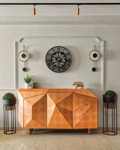 Faceted copper-toned console cabinet under a wall clock and sconces in a marble-floored foyer