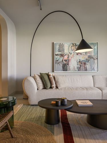 Beige sofa beneath abstract art, arc lamp overhead, dark round coffee tables on layered rug