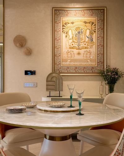 Round marble dining table with brass trim beneath a framed pichwai painting on a textured cream wall