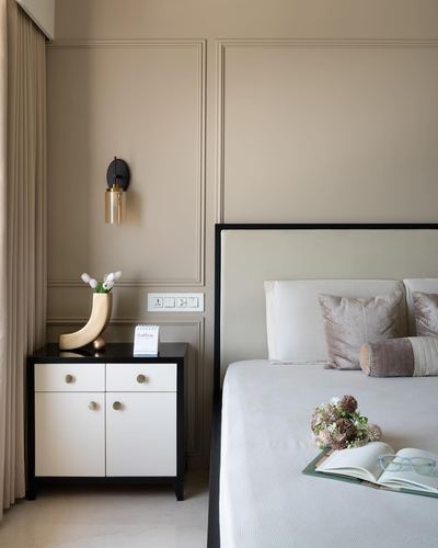 Beige paneled bedroom corner with black-and-white nightstand, wall sconce, and upholstered bed with pillows.