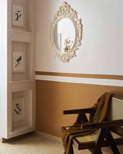 Ornate white mirror above a terracotta color-blocked wall beside a cane-webbed wooden chair with olive throw