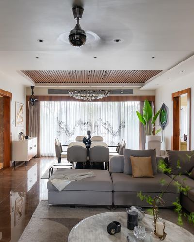 Open living-dining room with grey sectional, chandelier, and slatted wood ceiling above sheer-curtained windows