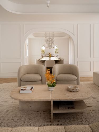 Cream living room with boucle armchairs and travertine table framed by arched doorway to dining room