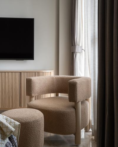 Curved boucle armchair and ottoman beside fluted wood sideboard near curtained window