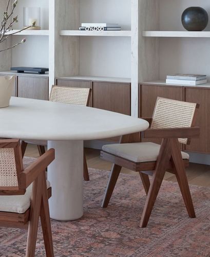 Oval stone dining table with cane-back wooden chairs before built-in plaster shelving and a patterned rug