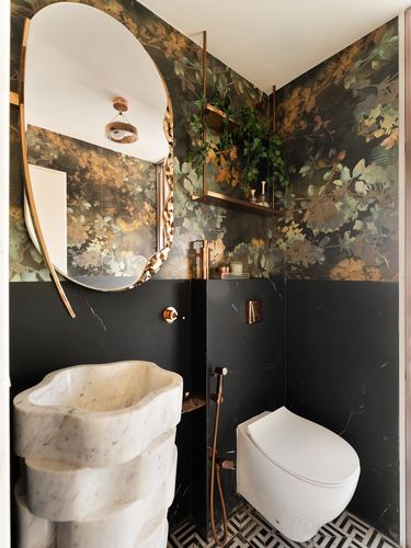 Powder room with floral dark wallpaper, oval brass mirror, carved marble pedestal sink and black marble walls