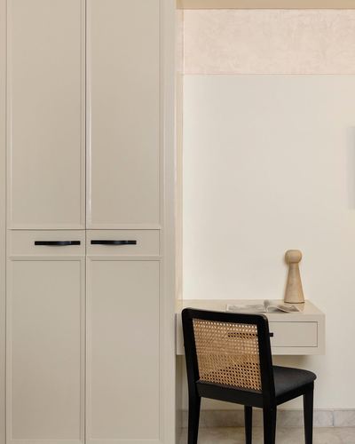 Floating desk with cane-backed black chair beside tall beige wardrobe with black handles