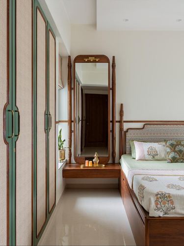 Narrow bedroom with green-trimmed cane wardrobe, standing wooden mirror, and floral upholstered bed.