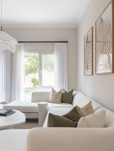 White living room with sectional sofa, layered pillows, framed prints, and garden view window