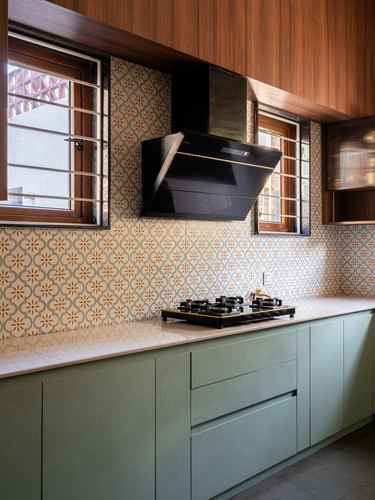 Kitchen counter with sage cabinets, patterned tile backsplash, black chimney hood between two windows