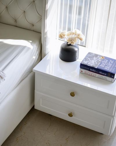 White glossy nightstand with dried flowers and stacked books beside a tufted headboard