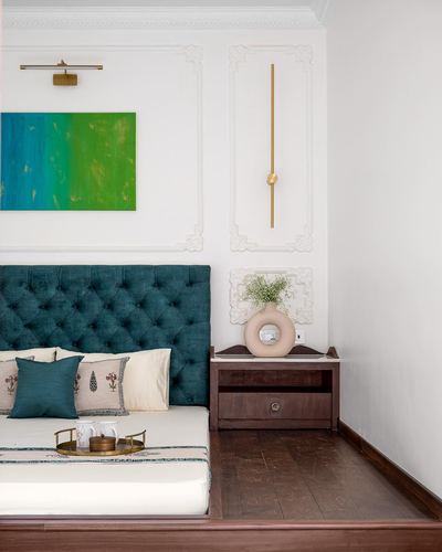 Bedroom corner with teal tufted headboard, molded white wall, wooden nightstand and donut-shaped vase